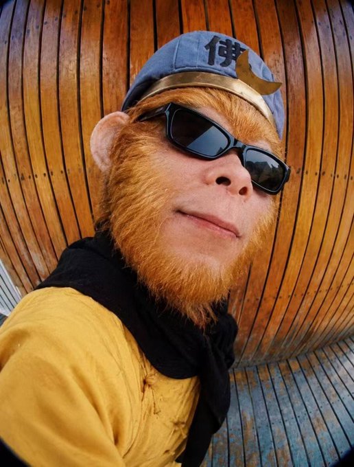 A person wearing a highly realistic monkey mask, black sunglasses, a blue headband with Chinese characters, a yellow garment, and a black scarf looks at the camera against a wooden slatted background.
