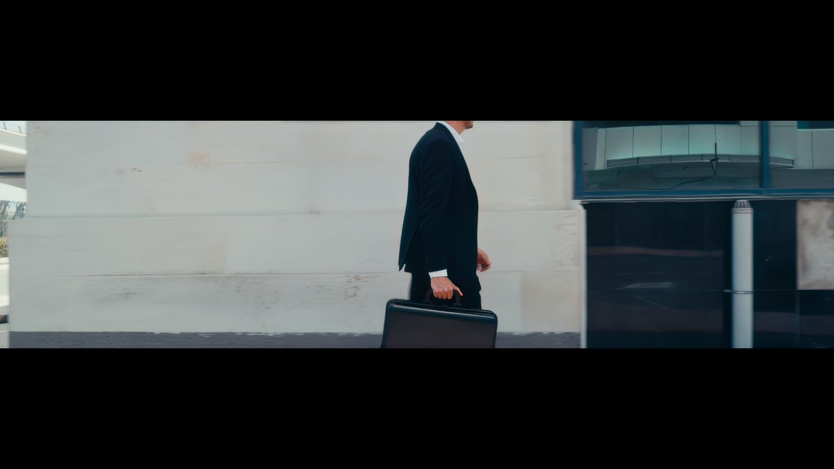 The image features a man dressed in a suit walking down a street while carrying a briefcase. The background includes a building with a glass window and a car parked nearby. The caption of the image reads, "New Wave x KAST We are beyond proud to be partnering with @KASTxyz as our first ever podcast sponsor! KAST is our neo-banking app of choice we use to spend stablecoins anywhere in the world, whilst earning rewards."