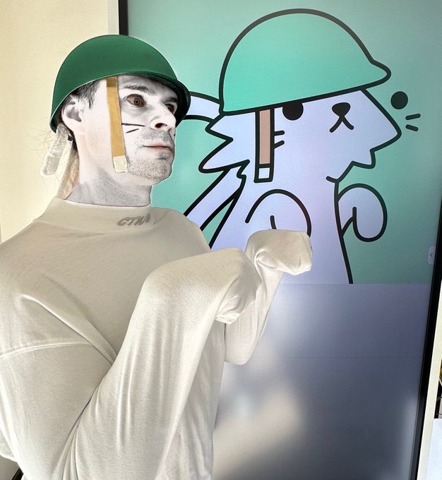 A person poses wearing a green military helmet with a chin strap and white face paint with drawn-on whiskers, standing next to a matching cartoon illustration of a cat in a green helmet.