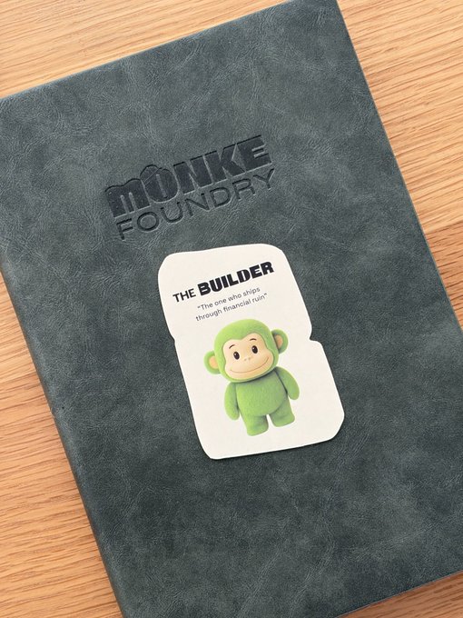 A black leather-bound notebook embossed with 'MONKE FOUNDRY' sits on a wooden surface, featuring a sticker of a green monkey character labeled 'THE BUILDER' with the quote 'The one who ships through financial ruin.'