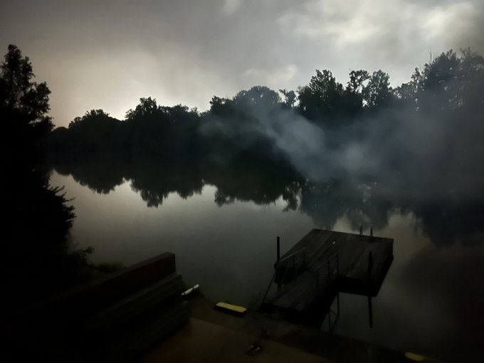 A misty, atmospheric view of a calm lake surrounded by dark silhouettes of trees, with a small wooden dock extending into the water in the foreground.