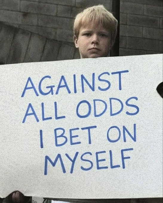 In the image, there's a young boy holding a sign that says " Against all odds I bet on my own". The boy appears to be standing against a brick wall with his hands on the sign. The text on the sign seems to be in a foreign language or written in an unusual style.