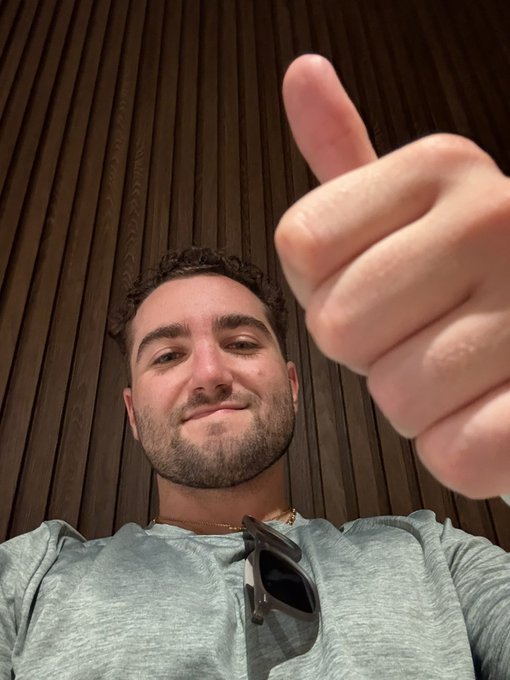 A young man with dark curly hair and a slight beard smiles while giving a thumbs-up, wearing a light gray shirt with sunglasses tucked into the collar, standing against a dark wooden slatted wall. The image conveys excitement and positivity, matching the post's announcement about his move to Miami to train and work as an attorney at the crypto/frontier tech law firm 0xhorizonslaw.