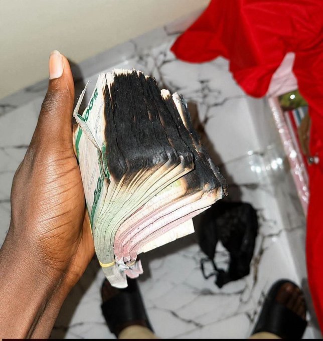 A hand holding a thick stack of Nigerian Naira currency notes that are heavily burned and blackened on the edges, with red fabric and a marble floor visible below.