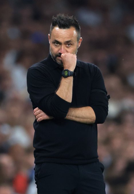 A man in a black long-sleeved shirt standing with his hand on his chin in a contemplative pose, wearing a smartwatch, against a blurred crowd background.
