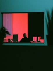 A silhouette of a person working on a laptop, illuminated by pink/red light coming through a window against a teal wall.