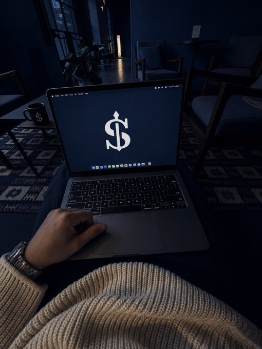 A person wearing a beige sweater sitting on a chair with a MacBook laptop on their lap displaying a dollar sign logo, in a dimly lit lounge area with a coffee mug on a side table.
