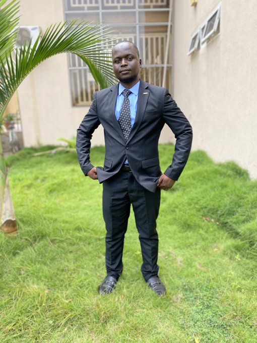 A man in a dark suit, blue shirt, and patterned tie posing on a grassy lawn in front of a building with palm fronds visible on the side.