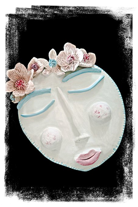 A decorative ceramic face mask adorned with pink flowers and turquoise closed-eye details against a black background.