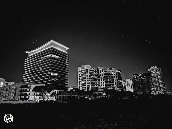 Black and white nighttime photograph of a city skyline with illuminated high-rise buildings and palm trees.