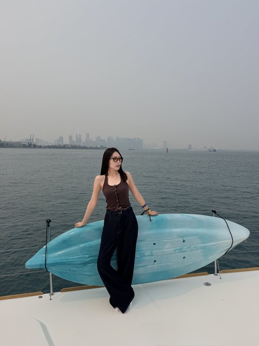 A woman in a brown tank top, black pants, and sunglasses stands on a boat holding a blue kayak, with a city skyline visible across the water in the background.