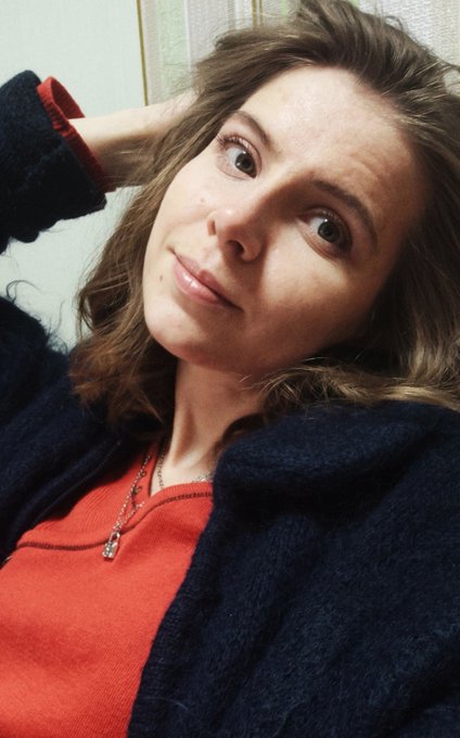 A close-up selfie of a woman with wavy brown hair wearing a red top and dark blue cardigan, looking at the camera with a slight smile.