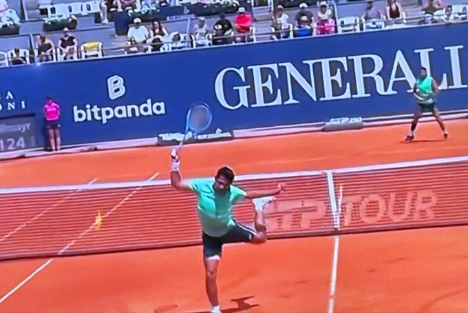 A tennis player in a teal shirt and white shorts serves on a clay court with 'GENERALI' and 'bitpanda' sponsor banners visible in the background.