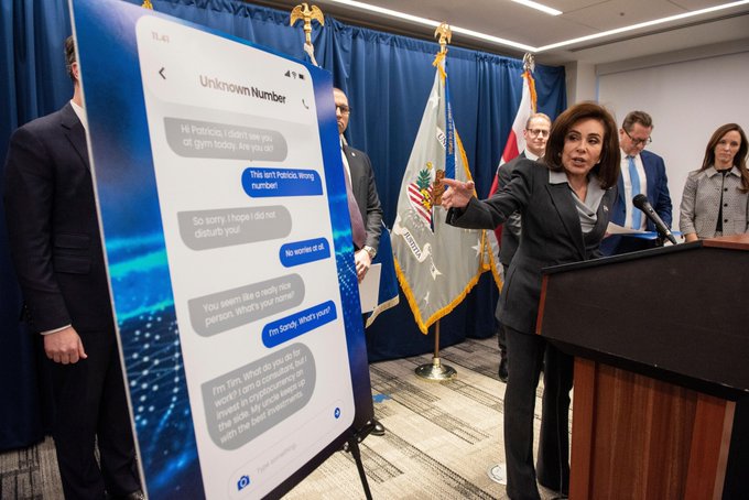 A press conference where a woman in a dark suit gestures toward a large smartphone display showing text messages from an 'Unknown Number', with official flags in the background.