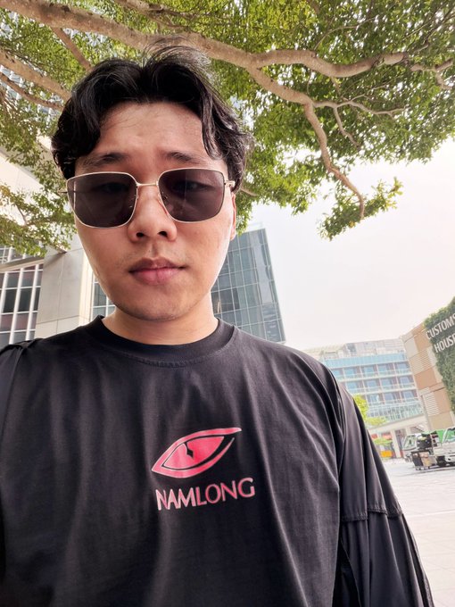 A selfie of a man wearing sunglasses and a black t-shirt with a red 'NAMLONG' logo, taken outdoors with trees and buildings in the background.