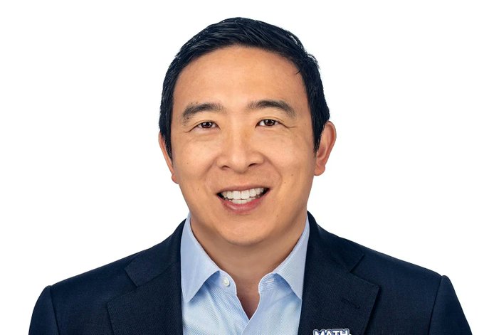 The image features a man wearing a suit jacket and tie with a blue shirt underneath. He has black hair and is smiling at the camera. The background of the image is white, which makes him stand out against it. There are no texts or other objects in the image, and the focus is solely on the man's face and attire.