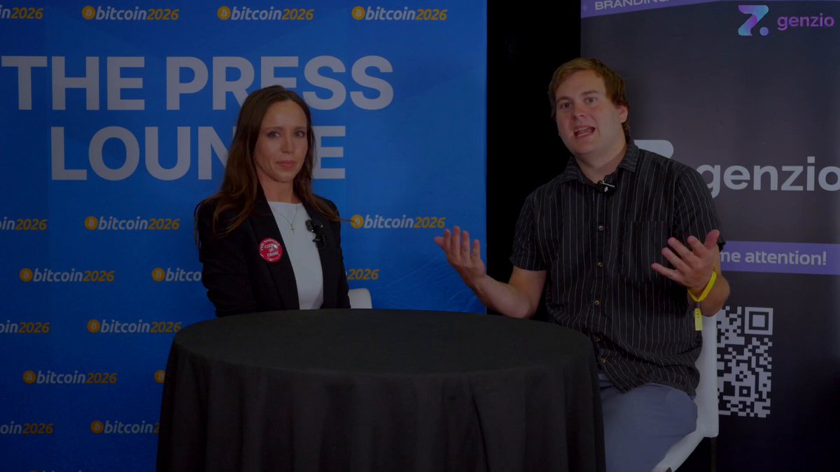 In this image, a man and woman are sitting across from each other at a table with a black tablecloth. They appear to be engaged in conversation or possibly participating in an interview. The background features a blue banner that reads "The Press Lounge" along with various logos such as Bitcoin, Genzio, and Dogecoin. There is also a Q&A format visible on the image, suggesting that they might be discussing a topic related to cryptocurrency or technology.