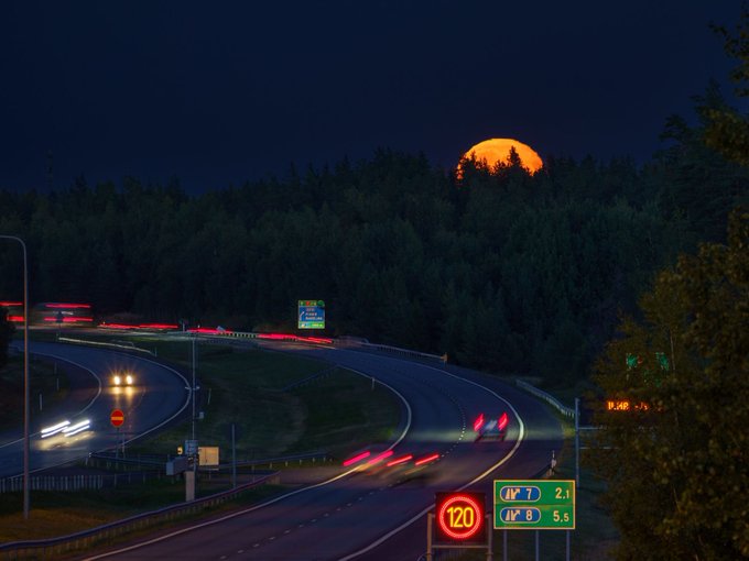 A nighttime time-lapse photo of a highway interchange shows streaking car lights, a glowing orange moon rising over the treeline, and a speed limit sign showing 120 km/h with route numbers 7 and 8.