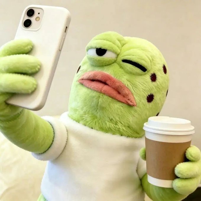 A green plush toy character holding a white smartphone in one hand and a coffee cup in the other, appearing to take a selfie.