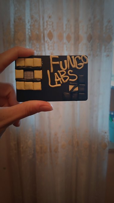 A hand holds up a dark blue card decorated with white chocolate chips and piped icing that reads 'FUNGO LABS' in a stylized font.