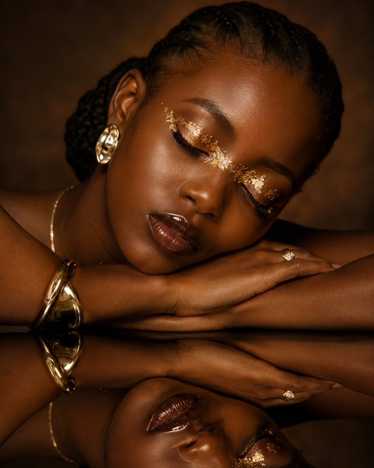 A beauty portrait of a woman with braided hair, gold earrings, and intricate gold face jewels, resting her head on her arms with a reflective surface below mirroring her features.