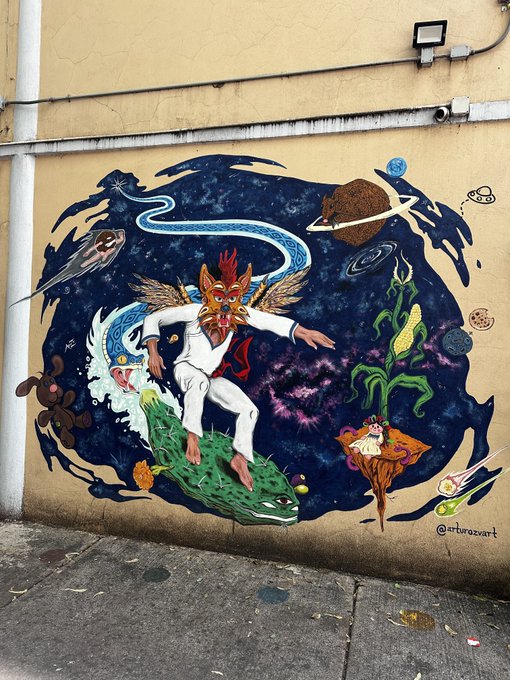 A vibrant street mural signed '@arturozvart' depicting a surreal cosmic scene with a masked figure riding a green crocodile-like creature through a starry galaxy filled with planets, a snake, and corn.