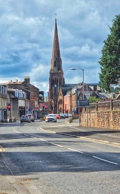 A street view of a town with a prominent tall church spire in the background under a cloudy blue sky.