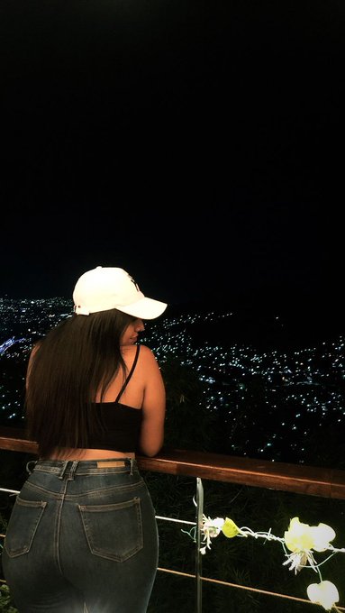The image features a woman standing on a balcony, looking out at a city skyline at night. She is wearing a black shirt and blue jeans, with her back turned towards the camera. The city lights create a beautiful backdrop for the scene as she gazes out over the view.