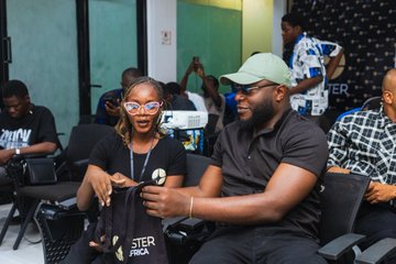 Two people, including a woman wearing a black shirt with "MASTER" visible, sit together at Aster's trading event in Lagos, engaged in conversation while other attendees gather in the background. The image captures the lively, community-focused atmosphere of the event described in the post, highlighting its role in guiding users toward trading success.