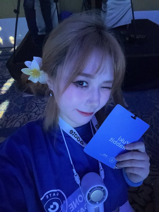 A young woman winks at the camera while holding up a blue "Gate.io" lanyard/badge, with another Gate lanyard draped around her neck featuring the iconic Gate logo, all bathed in the cool blue lighting characteristic of the Gate.io Summit on Day 2.