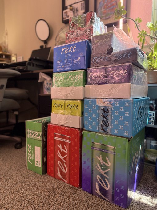 A tall stack of various colorful boxes branded with the logo 'rekt' sits on a carpeted floor in a room with a desk and plants.