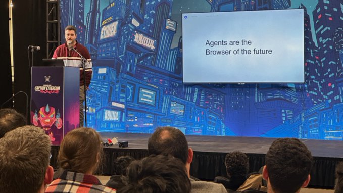 The image shows a man standing behind a podium giving a presentation, with an audience of people watching him intently from behind a curtain or screen. The man appears to be speaking about agents being the future of the browser industry. There are several people in the audience, some sitting and others standing, all focused on the speaker at his booth.