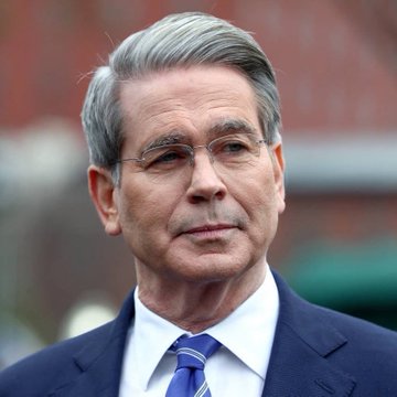 Treasury Secretary Scott Bessent is pictured in a close-up portrait, wearing a navy suit with a blue and purple striped tie, appearing outdoors. The image accompanies news that Bessent has accused Senate Democrats of blocking the Crypto Clarity Act.