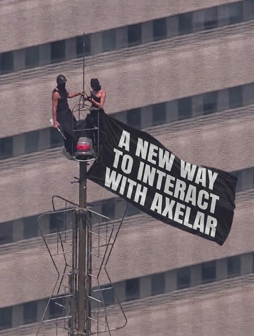 Two people on top of a communications tower or antenna structure unfurling a large black banner reading 'A NEW WAY TO INTERACT WITH AXELAR' against a backdrop of a tall building.