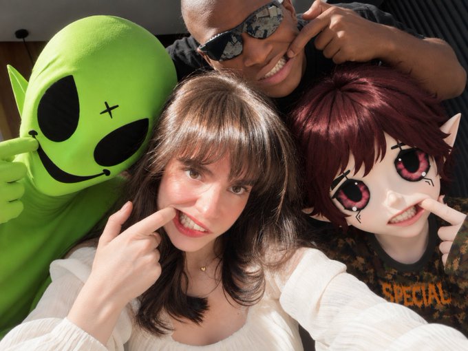 A selfie of three people playfully posing with their fingers near their mouths, including one wearing a green alien mask and another with an anime-style filter featuring large pink eyes and pointy ears.