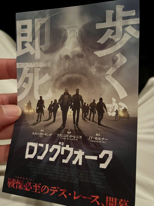 A hand holds a Japanese movie poster for 'Long Walk' featuring a face in the background and walking silhouettes, with text reading '即死ゲーム' and credits for stars including スカーレット・ヨハンソン and ザーン・マクグロー.