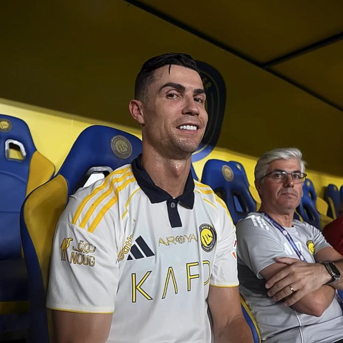 Cristiano Ronaldo sits in a stadium dugout wearing a white and yellow Al Nassr FC jersey, appearing alongside another man in a gray shirt.
