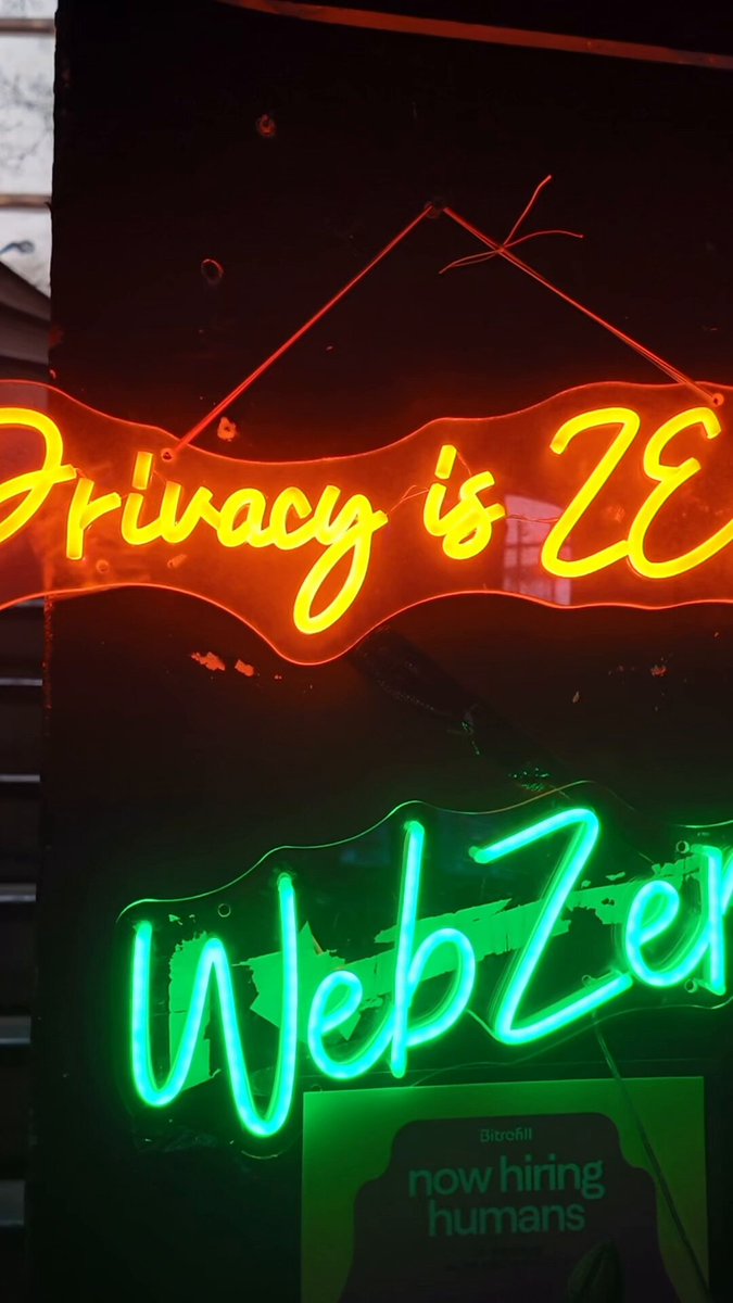 The image shows two neon signs hanging from a building, one orange and one green. Both signs are illuminated with white letters that say "WebZe" written in black cursive font. The background of the image is dark gray, which contrasts with the bright colors of the neon signs. The text on the signs reads "Inside of Blockspäti Berlin @joinwebzero @Zcash".