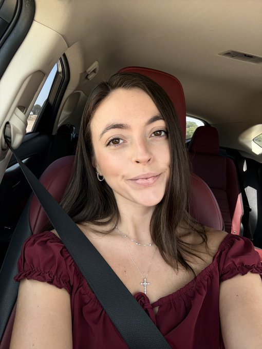 A selfie of a woman with long brown hair wearing a maroon off-shoulder top and a cross necklace, sitting in a car with a seatbelt on.