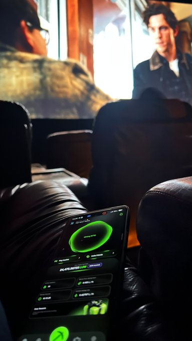 A person relaxes in a dark movie theater with Spider-Man playing on the screen in the background, while holding a phone in the foreground displaying the Lumira app's mining dashboard showing 24,475.92035 LUM earned, perfectly capturing the "watching SpiderMan, mining Lumira" multitasking moment highlighted in the post.