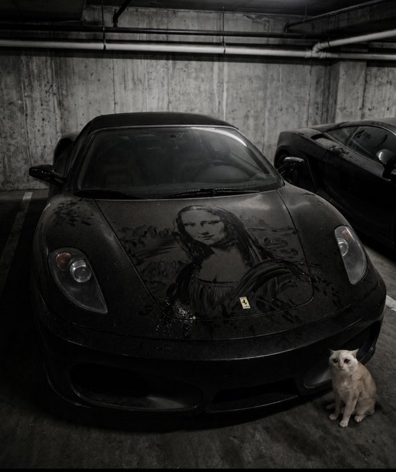 A black Ferrari sports car featuring a Mona Lisa design on its hood, parked in a concrete garage next to a light-colored cat.