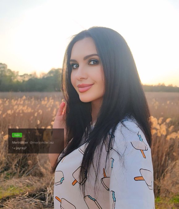 A woman with long dark hair in a patterned top stands in a field of tall grass at sunset, with a small overlay reading "MerinMiller @merinmiller.tez → jayray7".
