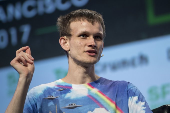 The image shows a man wearing a blue shirt with a rainbow design, standing behind a podium at an event. He appears to be giving a speech or presentation, as indicated by his hand gesture towards the camera. The background of the image features a large screen displaying text related to cryptocurrency and stablecoins, suggesting that this is likely a conference or gathering focused on the development and implementation of new ideas in the world of digital currency.