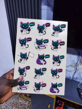 A hand holds up a sticker sheet featuring rows of a cartoon black and white cat character with pink and teal accents, accompanied by comic-style speech bubbles containing words like "DEV," "BYE," "EEEEK," "OMG," and "YEY!"—playful GenLayer branding that complements the post's message about the laptop repping the future of intelligent blockchains rather than a brand.