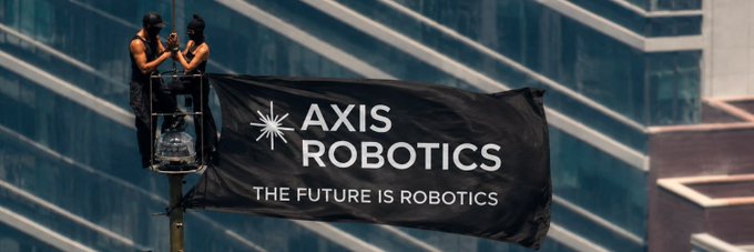 Two people on a lift installing a large black banner reading 'AXIS ROBOTICS - THE FUTURE IS ROBOTICS' with a starburst logo, set against the backdrop of a modern glass building.