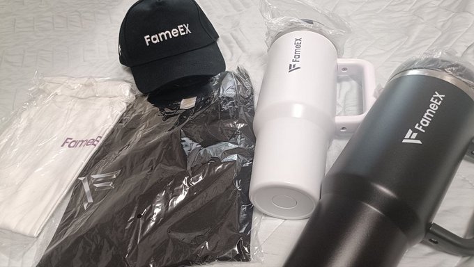 An overhead shot of branded merchandise including a black FameEX cap, white and black FameEX tumblers, and folded clothing items laid out on a white surface.