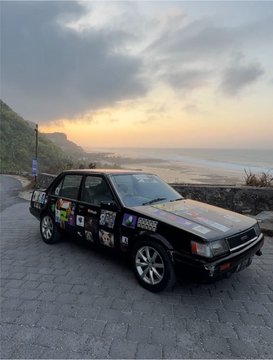 A black rally-style hatchback with sponsor decals including "#5555" is parked on a stone overlook at sunset, with a beach and ocean visible in the background.