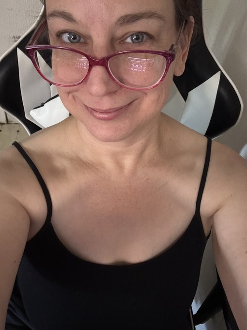 A selfie of a woman with red glasses, earrings, and a black top smiling at the camera.