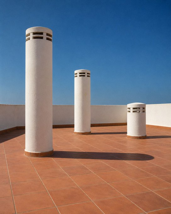 Three white cylindrical ventilation pipes of varying heights stand on a terracotta-tiled rooftop against a clear blue sky.