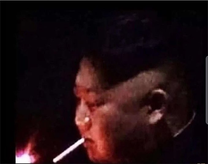 A dark, blurry photo of a man's profile in low light, appearing to smoke with a small flame visible in the corner.